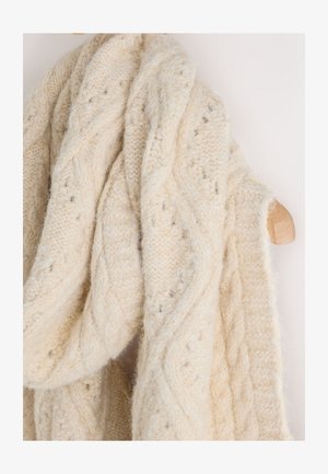 Šal krem boje, pleten grubom tehnikom s uzorkom pletenica i tekstura, prebačen preko drvenog vješalice na svijetloj pozadini.