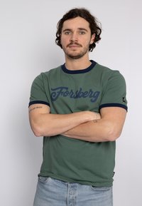 Camiseta de algodón verde con cuello y puños de canalé en navy. Presenta un audaz gráfico azul de "Forsberg" en la parte frontal. Ajuste casual, mangas cortas.
