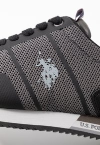 Baskets en maille noir et gris avec logo en relief et accents texturés. Présente une semelle en caoutchouc blanche et un système de laçage noir.