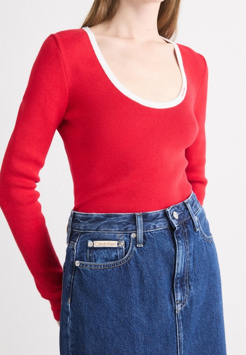 Personne portant un haut rouge à manches longues avec un col rond et une bordure blanche, ainsi qu'une jupe en jean bleu taille haute Calvin Klein.
