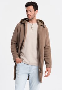 Brun hættejakke med lynlås, som har en struktureret stof, hvide knapper og en længere længde. Båret over en lys beige top.