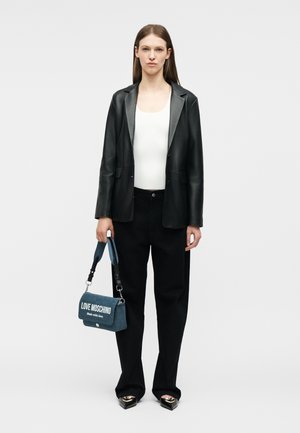 Γυναίκα που φοράει μαύρο δερμάτινο σακάκι, λευκό τοπ, μαύρο παντελόνι, κρατώντας μια μπλε τσάντα Love Moschino, στέκεται μπροστά από ένα απλό λευκό φόντο.