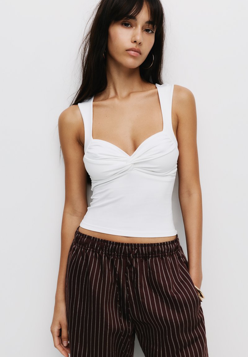 Hvid cropped top med et snoet knudedetalje foran, brede skulderstropper, parret med brune stribede løstsiddende bukser. Glat stofstruktur.