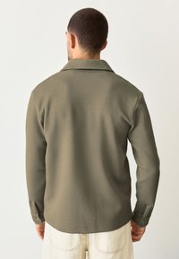 Camicia texture verde oliva con colletto, maniche lunghe e polsini bottonati. Presenta un motivo a coste diagonali e una vestibilità rilassata.