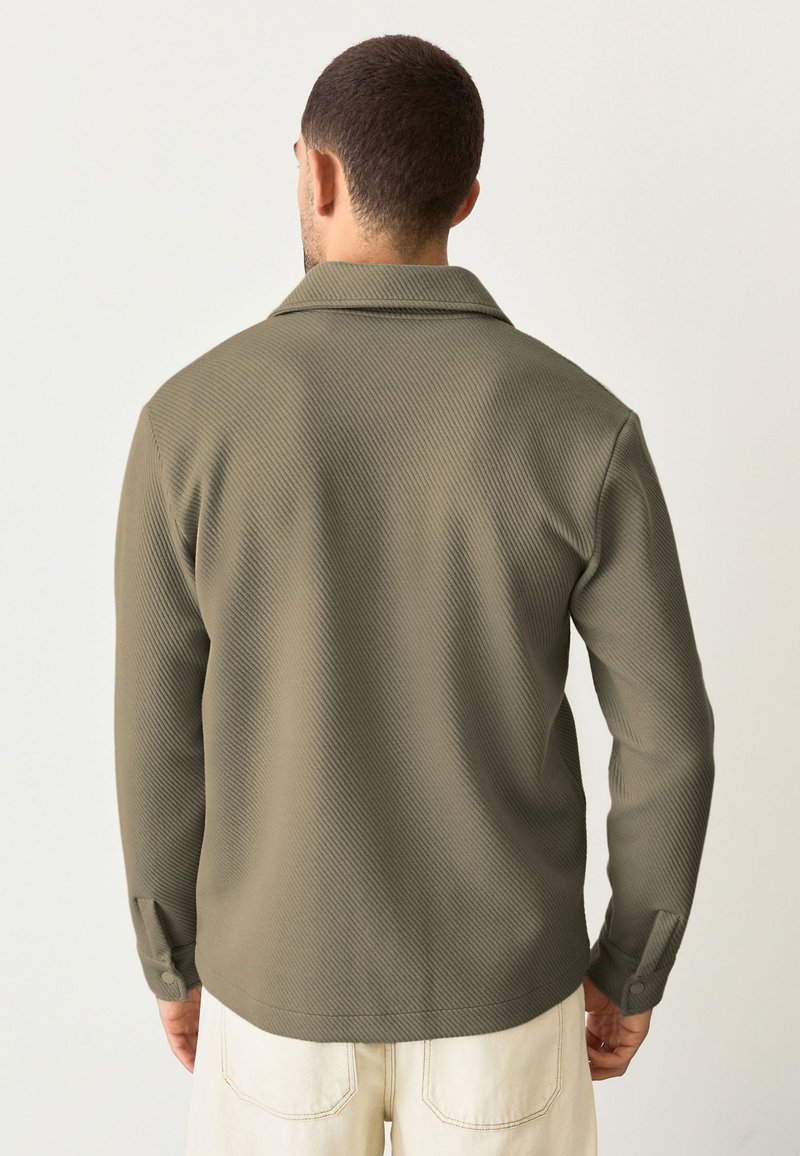 Camicia texture verde oliva con colletto, maniche lunghe e polsini bottonati. Presenta un motivo a coste diagonali e una vestibilità rilassata.