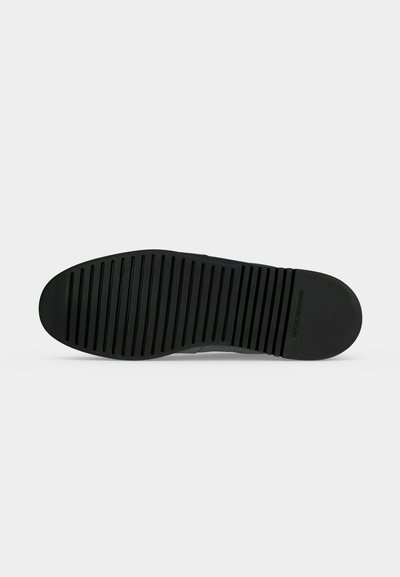 Semelle de chaussure noire avec des rainures horizontales pour l’adhérence et le nom de la marque en relief près du talon, présentée sur un fond clair uni.