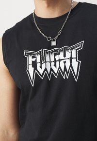 Detailní záběr na osobu v bezrukávovém černém tričku s nápisem "FLIGHT" napsaným b�ílým zubatým písmem a stříbrným náhrdelníkem s přívěskem ve tvaru zámku.