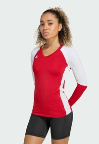 Maglietta atletica a maniche lunghe rossa e bianca con scollo a V, abbinata a pantaloni corti neri. La texture del tessuto appare liscia e aderente.