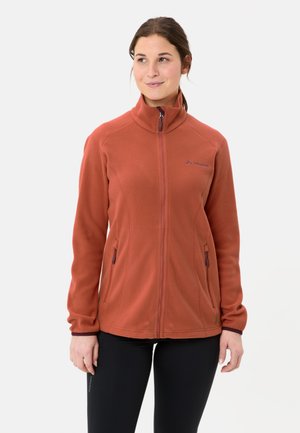 Donna che indossa una giacca in pile color ruggine con cerniera e colletto alto, pantaloni neri, in piedi davanti a uno sfondo chiaro e uniforme.