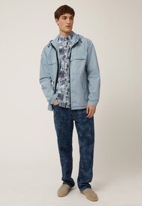 Chaqueta con capucha de color azul claro con dos bolsillos delanteros, una camisa de patrón azul y blanca debajo, y pantalones oscuros de textura azul. Zapatillas color beige sin cordones.
