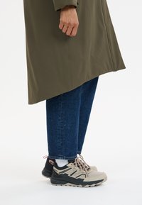 Olijfgroene lange jas gecombineerd met blauwe denim jeans. Grijs en zwarte sneakers met rode accenten, platte veters en gevoerde zolen.