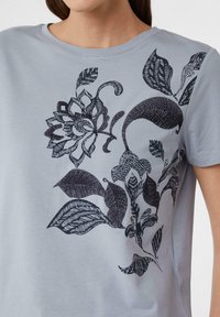 Błękitny T-shirt z czarnym wzorem kwiatowym i różnymi wzorami liści. Materiał wydaje się gładki, z okrągłym dekoltem.