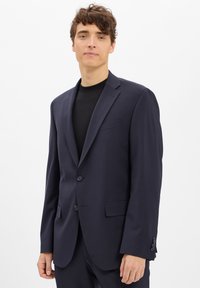 Granatowy blazer wykonany z gładkiego materiału, z dwoma guzikami, pojedynczą kieszenią na piersi i dopasowanym krojem. Noszony na czarnej koszuli.