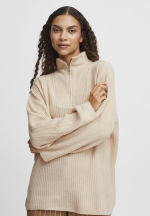 Beiger, gerippter Reißverschluss-Pullover in weicher Textur, mit hohem Kragen und lockerer Passform. Auffälliges silbernes Reißverschlussdetail am Hals.