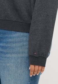 Grå sweatshirt med ribbade muddar, bärs över blå jeans. Har en liten logotypaccent på mudden. Slät textur, avslappnad passform.