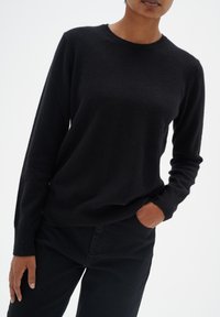 Schwarzer Strickpullover mit langen Ärmeln, runder Ausschnitt und weicher Textur. Getragen mit schwarzen Hosen, die Hand in der Tasche, was einen lässigen Look zeigt.
