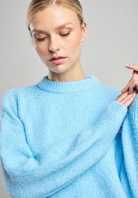 Pull en tricot bleu clair avec une texture côtelée, un col rond et des manches longues. Le tissu semble doux et duveteux.