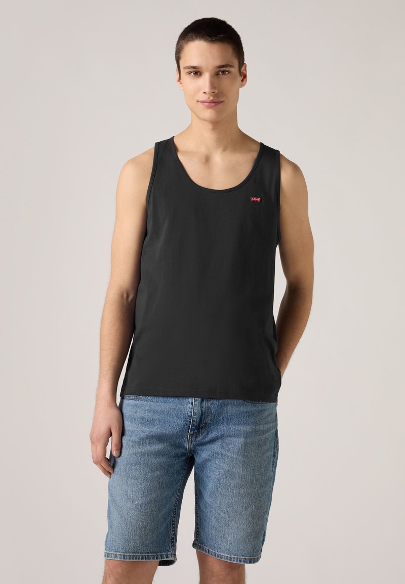 Junger Mann, der ein schwarzes ärmelloses Tanktop und blaue Jeansshorts trägt, steht vor einem einfarbigen beigen Hintergrund.