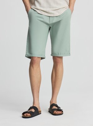 Uomo con pantaloncini verde chiaro con orlo arrotolato e sandali neri con fibbia, in piedi con le mani in tasca davanti a uno sfondo semplice.