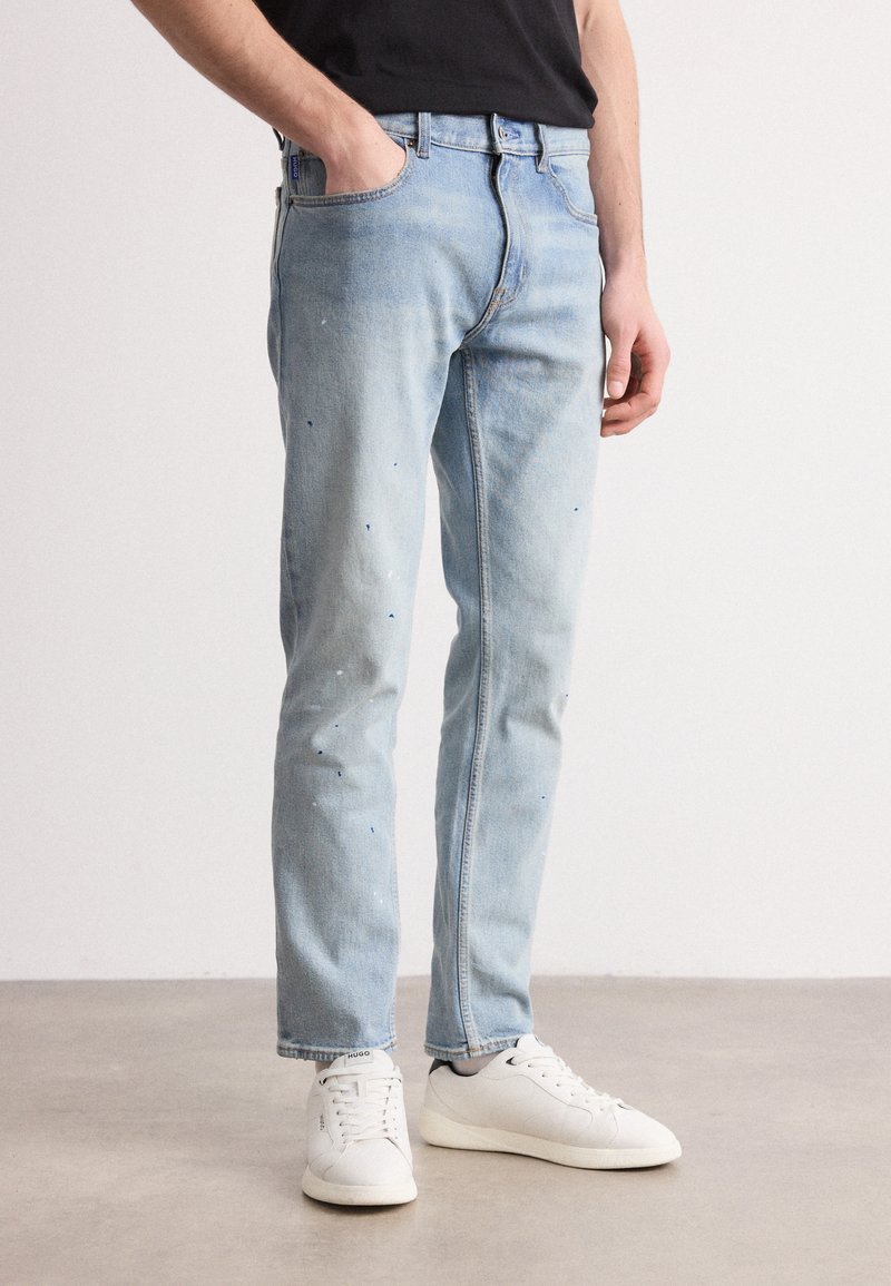 Jeans de mezclilla azul claro con corte recto, exhibiendo acentos de salpicaduras de pintura. Combinados con zapatillas blancas. Cintura sencilla y diseño clásico.