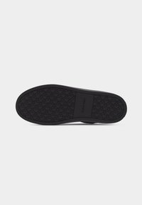 Semelle de chaussure noire avec motif de bande de roulement et logo "Calvin Klein" imprimé au centre, sur un fond uni gris clair.