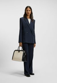 Completo doppiopetto blu navy con rever ampi, abbinato a una borsa strutturata bicolore con base nera e pannello frontale crema.