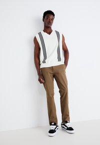 White sleeveless sweater with black vertical stripes, paired with light brown pants and black and white sneakers, standing against a plain backdrop.