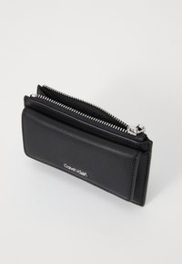 Svart läderplånbok med dragkedjestängning, slank design och en silverfärgad Calvin Klein-logotyp på framsidan. Slät struktur med metallakcenter.