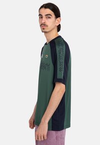 Camiseta deportiva de manga corta verde con acentos en navy, que presenta un patrón geométrico en los hombros y un logo en el pecho. Textura de tela suave.