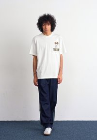 Hvid bomuld T-shirt med trykt grafik på venstre bryst; parret med marineblå træningsbukser med sidelister og hvide sneakers.