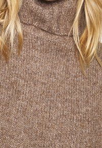 Pull à col roulé en tricot marron présentant une texture douce et une coupe ample, avec des motifs de points visibles et de subtiles variations de couleur.