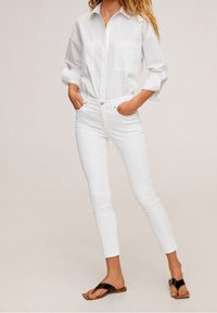 Camisa blanca de botones con mangas enrolladas, combinada con jeans blancos ajustados. Sandalias planas marrón y negras completan el atuendo.