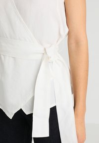 Top bianco avvolgente realizzato in tessuto leggero, con nodo laterale, orlo asimmetrico e una texture liscia. Abbinato a jeans scuri.