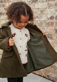 Olivengrøn uldfrakke med tekstureret overflade, med plaid-foring og lomme detaljer, båret over en cremefarvet sweater med blomsterbroderi.
