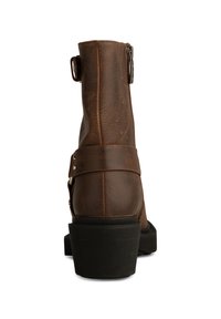 Bota de tobillo de imitación de cuero marrón con cremallera lateral, detalle de hebilla ajustable y suela de goma texturizada. Presenta un tacón bajo apilado.
