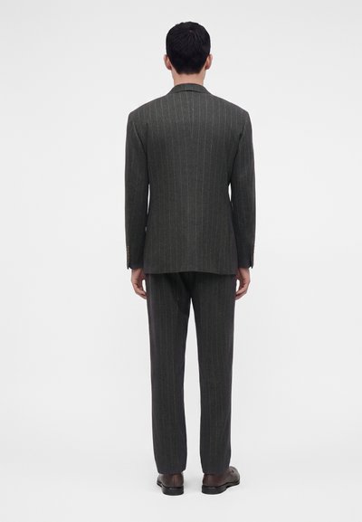 Homme debout, tourné dos à la caméra, portant un costume gris foncé à fines rayures avec un pantalon assorti et des chaussures marron sur un fond uni.