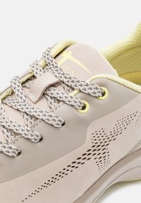 Jasno beżowe buty sportowe z gładką skórzaną cholewką, siateczkowym wnętrzem, jasnymi żółtymi akcentami i szarymi teksturowanymi sznurowadłami. Perforowany wzór po bokach.