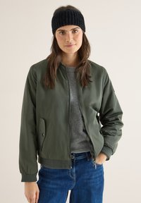 Chaqueta bomber verde con una textura suave y brillante, puños acanalados y cierre delantero con cremallera. Llevada sobre un suéter gris con vaqueros azules. Accesorio de diadema negra.