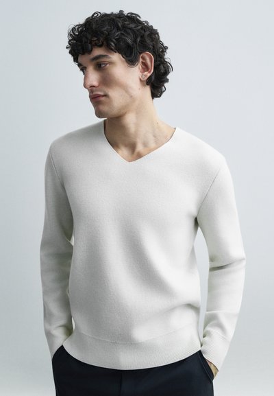 Weißer V-Ausschnitt-Pullover aus leichtem Strick mit langen Ärmeln, der einen gerippten Saum und Bündchen aufweist. Das Modell steht vor einem hellen Hintergrund.