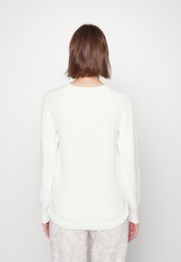 Pull de couleur crème avec une texture côtelée, manches raglan et ourlet arrondi. Porté avec un pantalon clair à motifs.