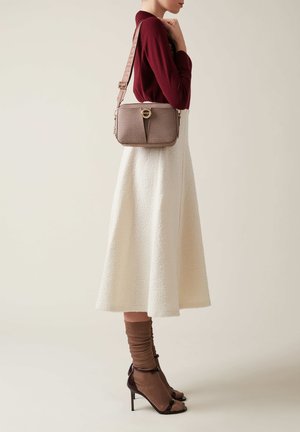 Donna che indossa un top bordeaux e una gonna midi in crema con texture, mentre porta una borsa a tracolla in taupe con una larga tracolla e una chiusura circolare.