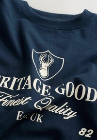 Tumesinine pusa, millel on uluki pea graafika ja valge tekst "Heritage Goods", ümara kaelusega, pehme tekstuuriga.