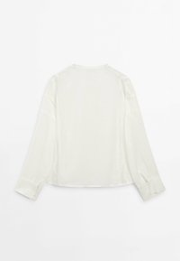 Blouse blanche à manches longues avec poignets boutonnés, ourlet droit et détails subtils de coutures, posée à plat sur un fond blanc.