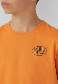 Oranje katoenen T-shirt met ronde hals. Bevat zwarte "PARADISE" tekst in een ovale vorm aan de linkerkant. Soepele textuur.