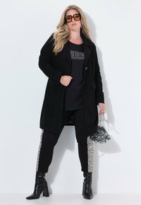 Cappotto lungo in corda nera, abbinato a un top nero con accenti di strass, leggings neri con pannelli a stampa leopardata e stivali con tacco.