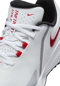 Pantofi de golf albi cu material texturat, accente negre și roșii, design perforat și șireturi albe cu logo-ul roșu Nike.