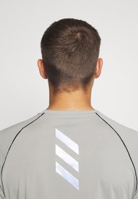 Šedé sportovní tričko s texturovanou tkaninou, krátkými rukávy a reflexním logem se třemi pruhy na horní části zad, zvýrazněné černými švy.