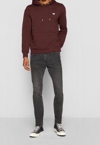 Burgundy hoodie med en framficka, snörning i huvan och en liten logotyp. Bärs med mörkgrå jeans och svarta låga sneakers med vita sulor.