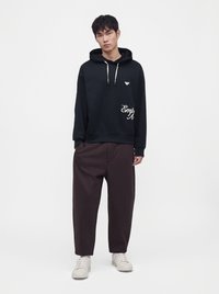 Jeune homme portant un sweat à capuche noir avec des cordons blancs, un pantalon sombre et ample, et des baskets blanches, debout devant un fond uni.