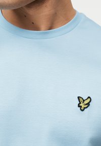 Κοντινό πλάνο ενός ατόμου που φοράει γαλάζιο μπλουζάκι με στρογγυλή λαιμόκοψη και ένα μικρό κίτρινο πουλί κεντημένο στο στήθος.